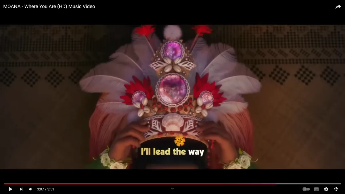 A close-up shot from the 'Moana' music video shows a character wearing an ornate, feathered headdress with red and white…