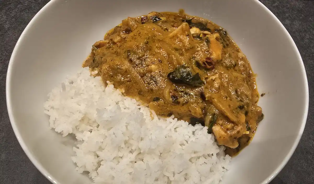 A white bowl contains a serving of white rice next to a portion of yellowish-brown curry with visible pieces of meat and…