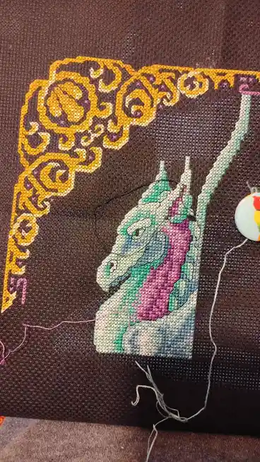 A close-up of a cross-stitch project on dark brown fabric, showing a partially stitched unicorn head with a purple mane and…