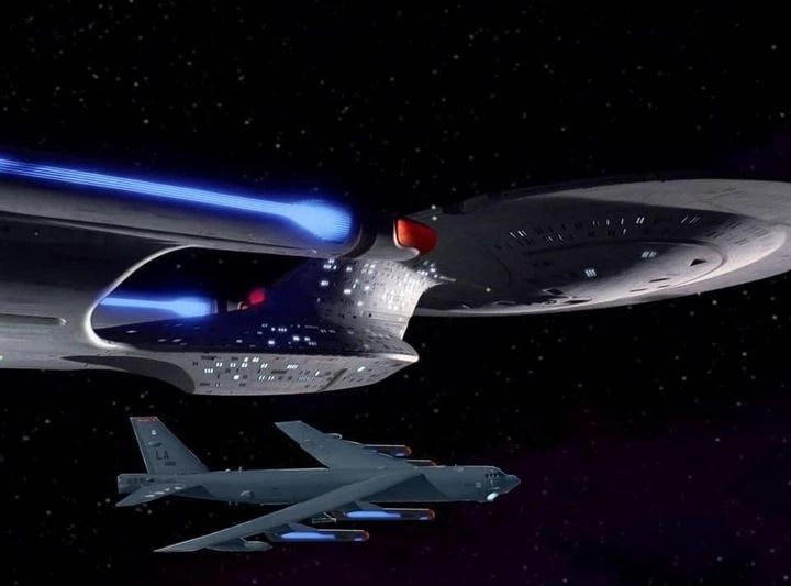 A digitally manipulated image shows a gray B-52 Stratofortress bomber flying in space beneath a large, silver Starfleet…