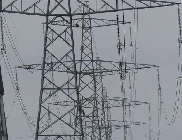 A photograph of a large metal electricity pylon with wires and insulators, against a gray sky. Two small figures are visible…
