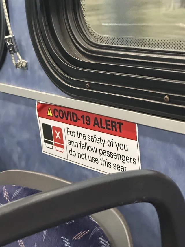 A close-up photo of a red and white COVID-19 alert sticker on a public transit seat, showing a black square with an X over a…
