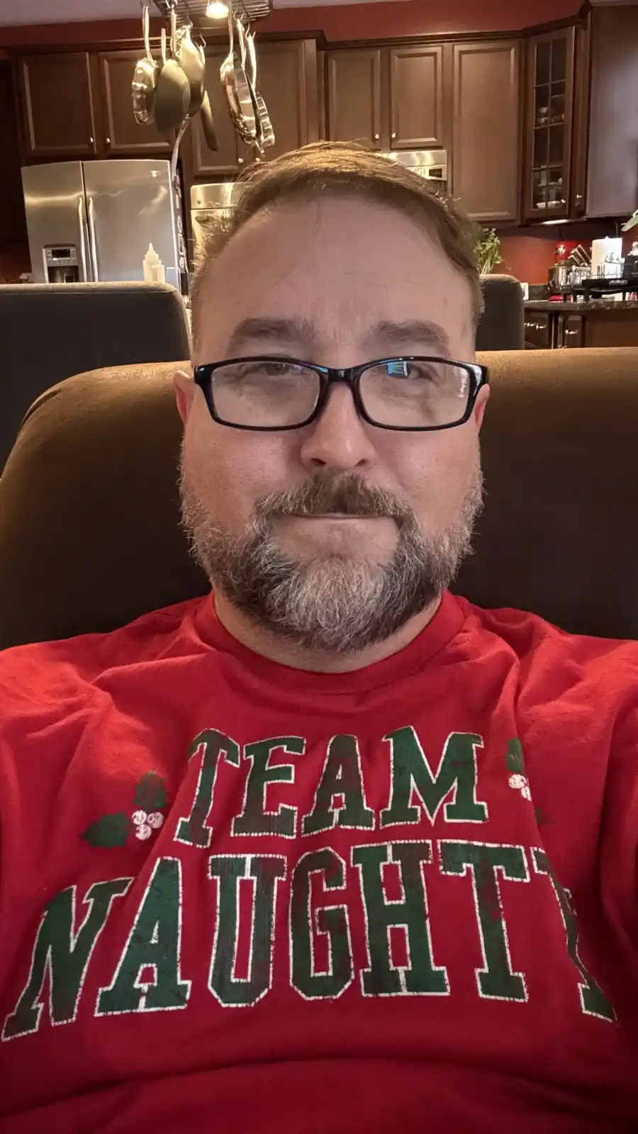 A man with glasses and a graying beard sits in a brown chair wearing a red t-shirt that reads 'TEAM NAUGHTY' in green…