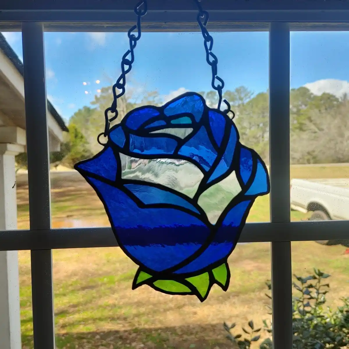 A blue stained glass rose ornament hangs by chains from a window with black mullions, showing a suburban yard and sky…