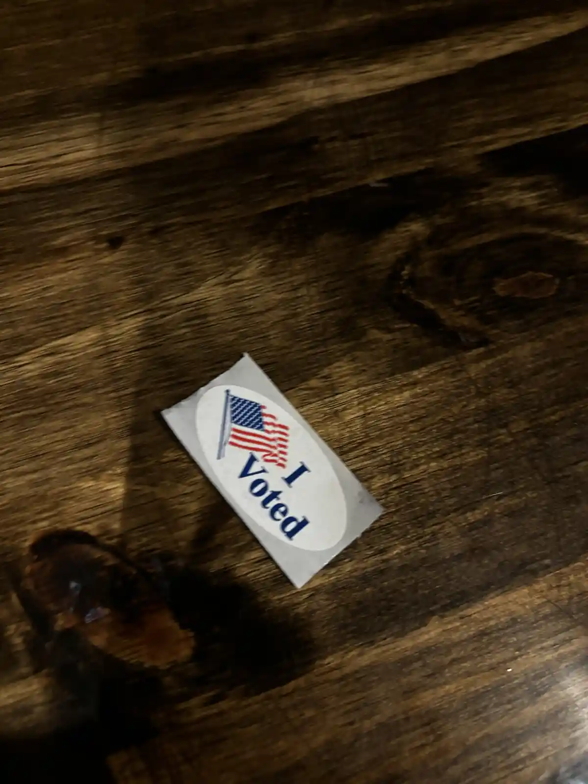 A close-up photo shows a white oval 'I Voted' sticker with a small American flag graphic, lying on a dark brown wooden…
