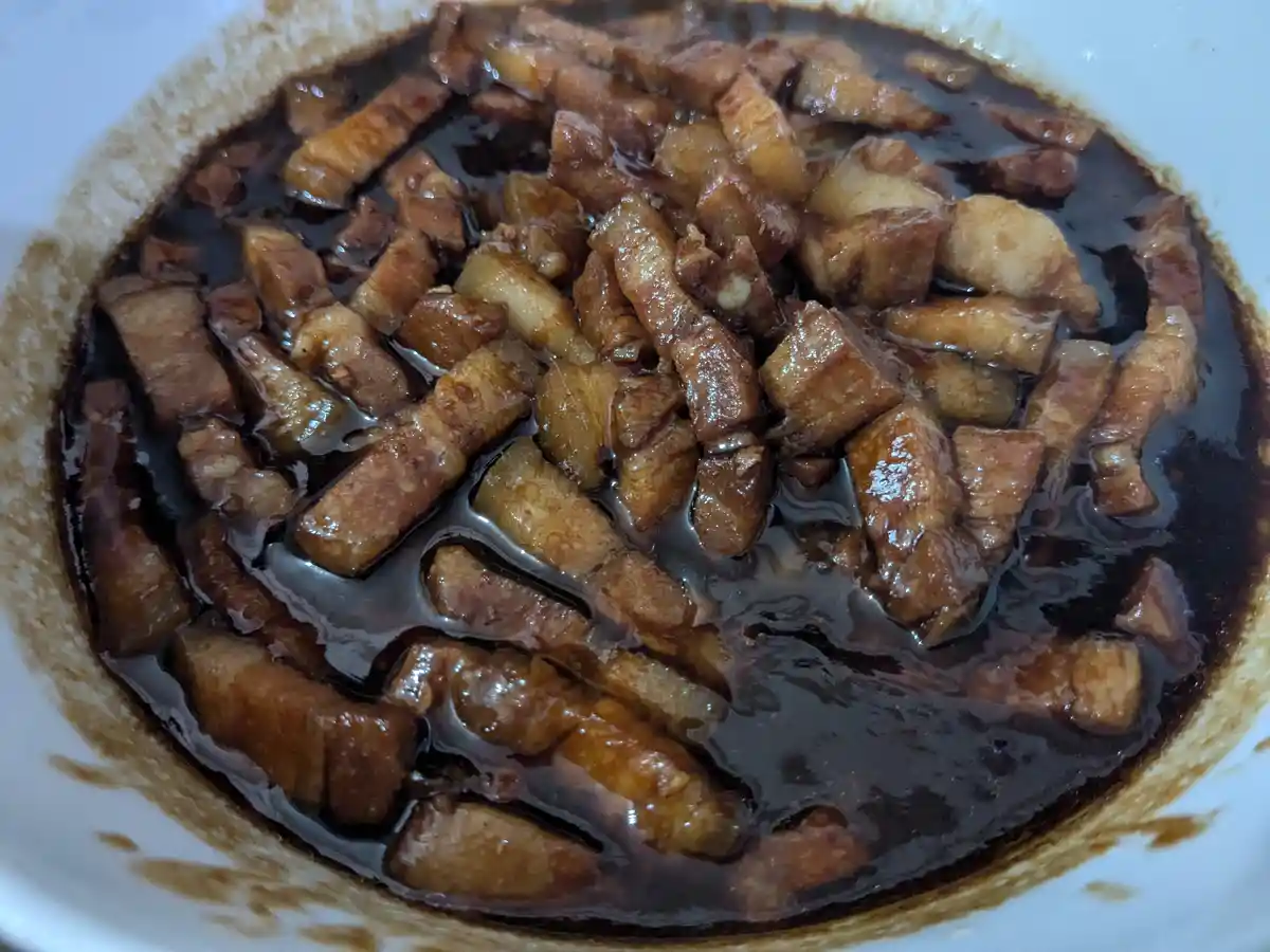 A close-up photo shows a white bowl filled with glossy, dark-brown braised pork belly pieces submerged in a thick, dark…