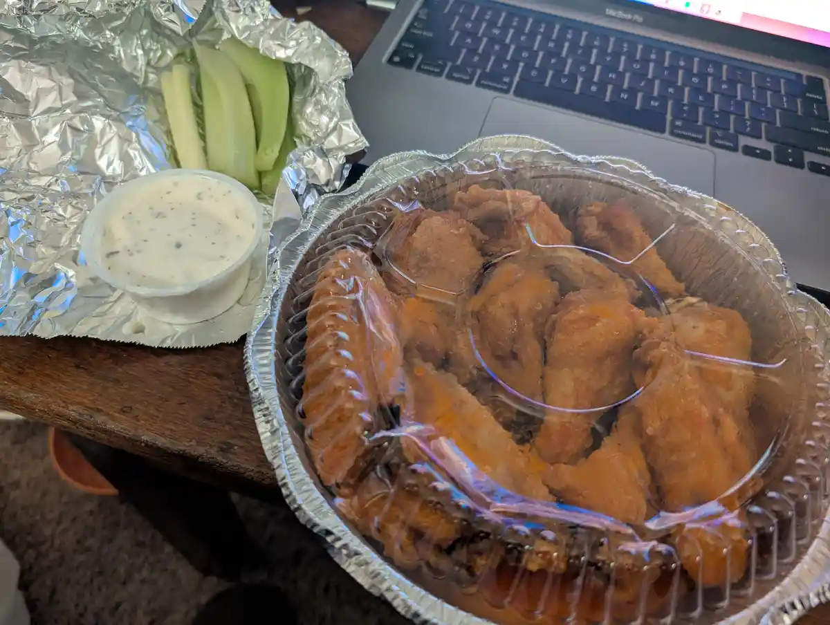 A close-up photo shows a takeout container of buffalo wings with orange sauce, accompanied by celery sticks and ranch…