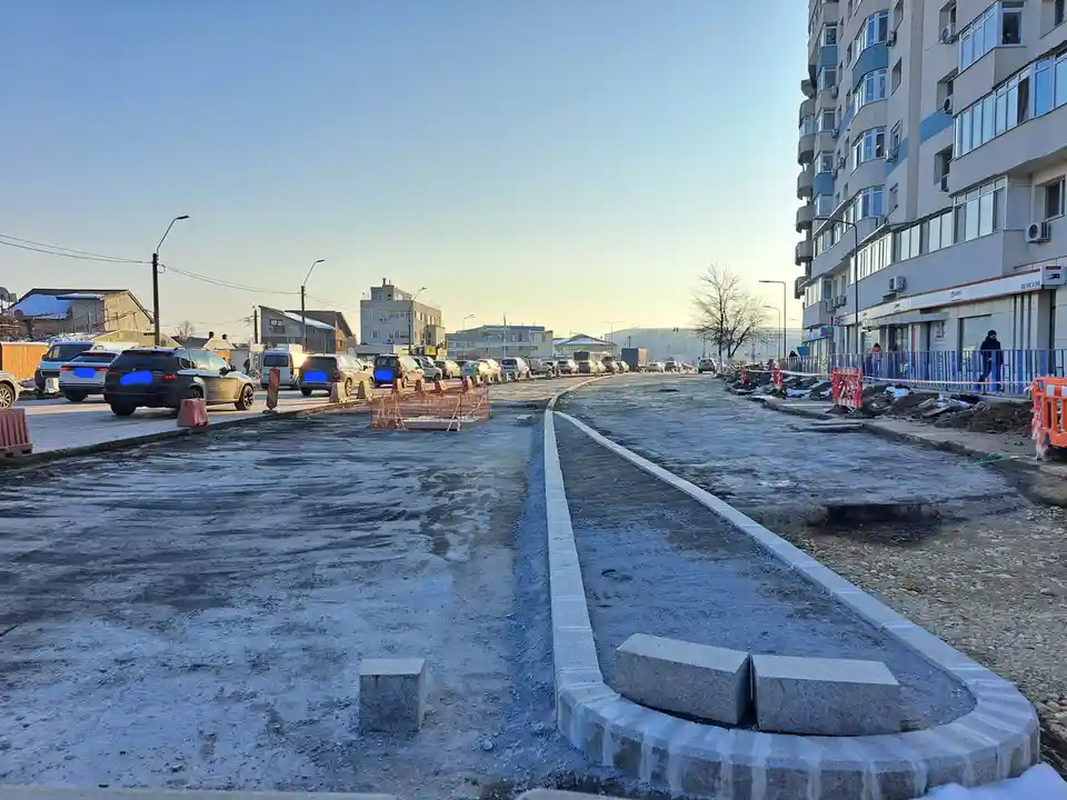 A street under construction with partially paved asphalt, concrete curbs, and orange safety barriers, cars parked on the…
