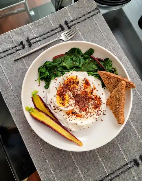 A top-down photo of a white plate with a healthy breakfast: sliced hard-boiled eggs on yogurt, spinach, two slices of whole…