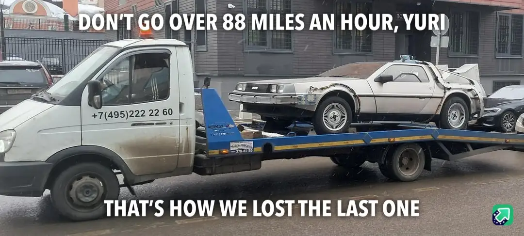 A white tow truck with a blue flatbed carries a damaged white DeLorean DMC-12, referencing the 'Back to the Future' movie…