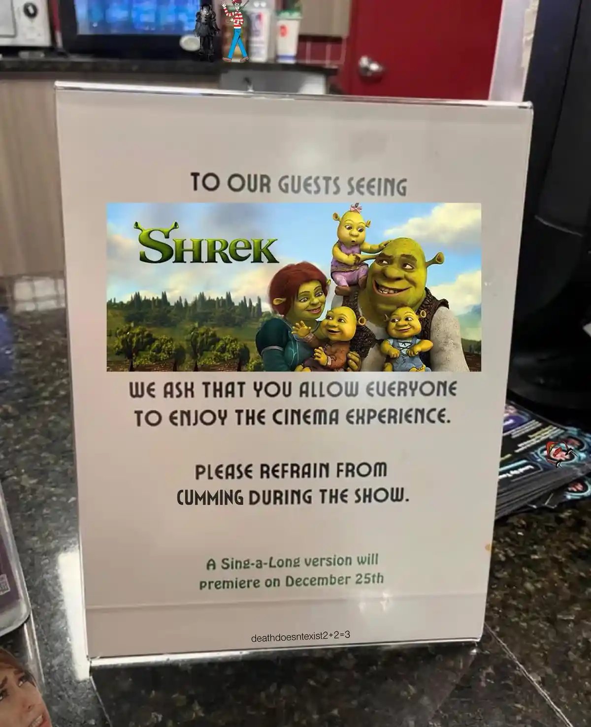 A sign for a movie theater showing 'Shrek' features a family photo from the film. The sign politely asks guests to allow…