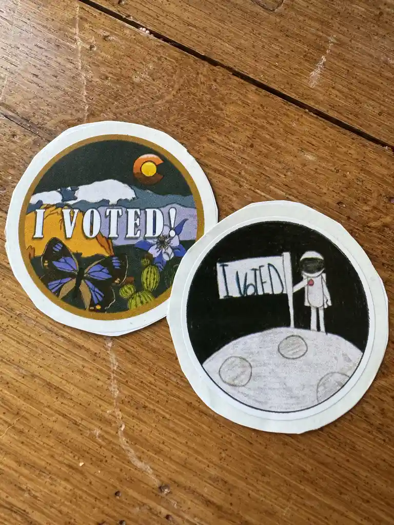 Two circular 'I VOTED' stickers lie on a wooden surface. The left sticker features a colorful, official-looking design with…