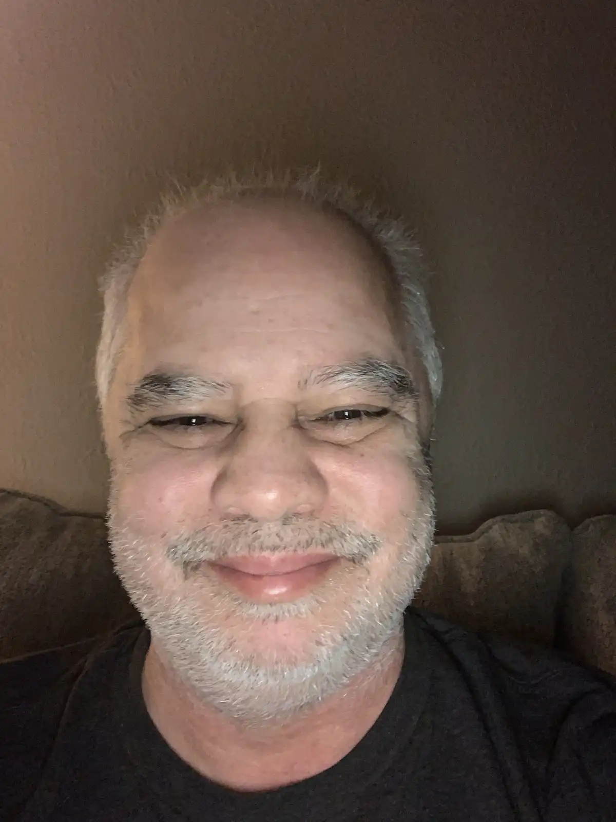 A close-up selfie of a middle-aged to older man with a balding head, gray hair, and a gray beard, smiling with squinted eyes…