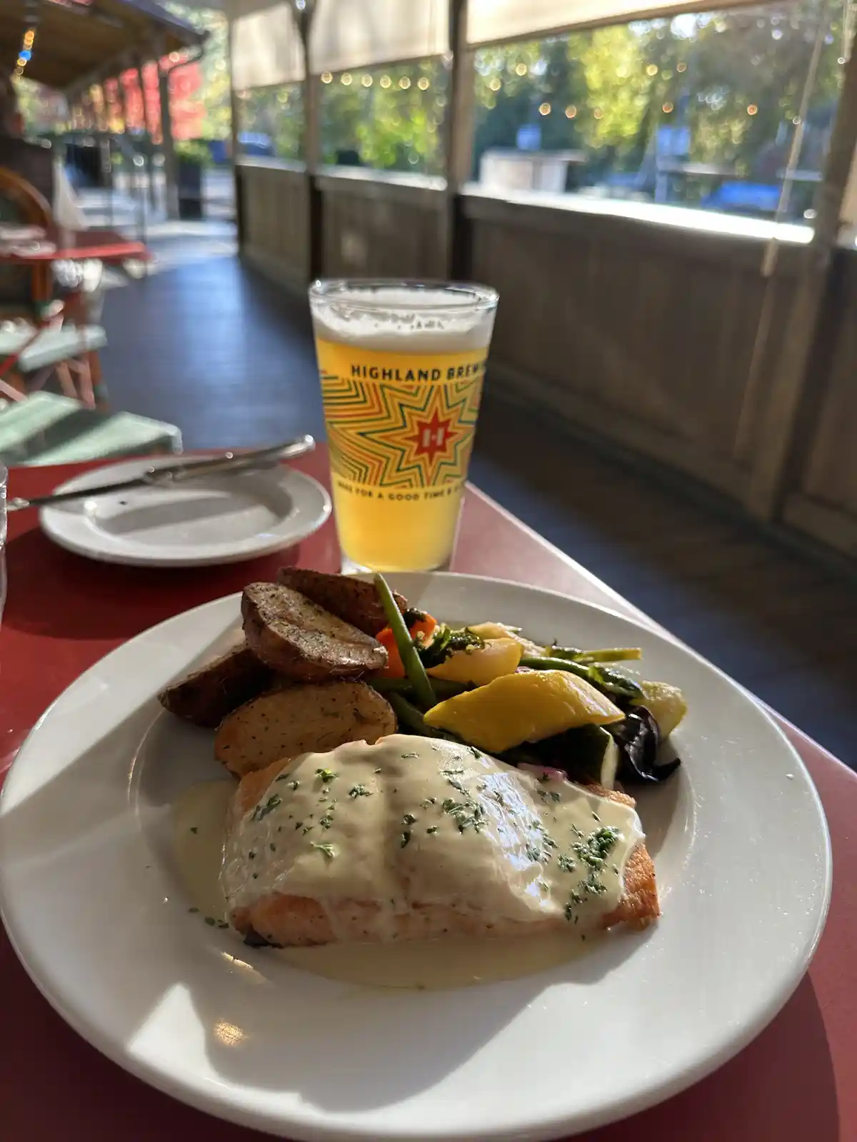 A plate of salmon with creamy sauce and roasted potatoes sits on a red table next to a glass of beer from Highland Brewing…