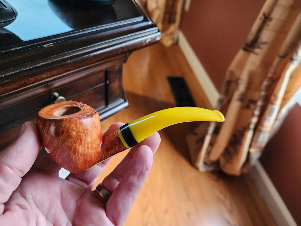 A hand holds a wooden smoking pipe with a bright yellow, curved mouthpiece, which is the focal point. The pipe has a dark…