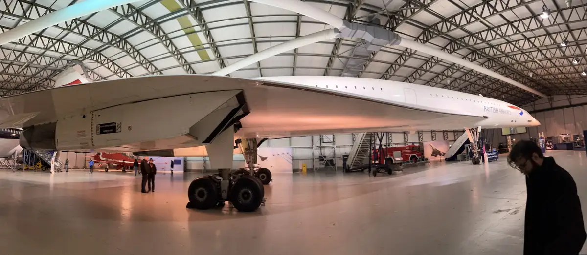 A man with glasses and a beard looks downcast in the foreground of a large hangar, while a massive white British Airways…