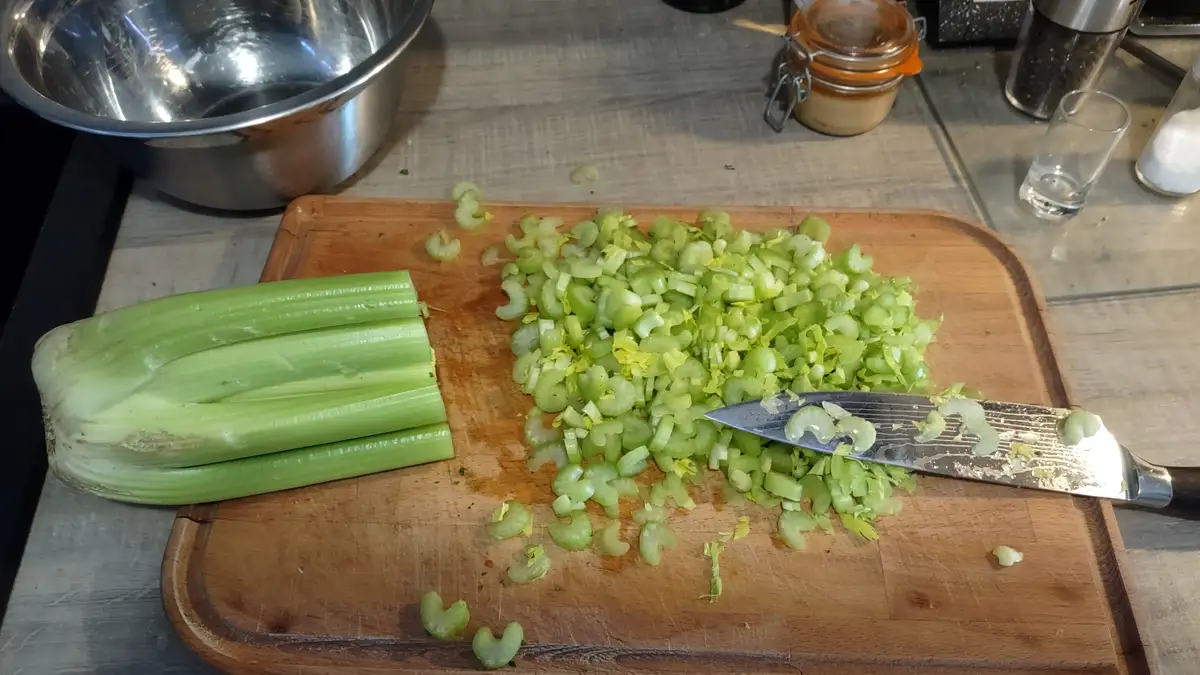A wooden cutting board holds a pile of freshly chopped green celery next to three unchopped celery stalks, with a knife…