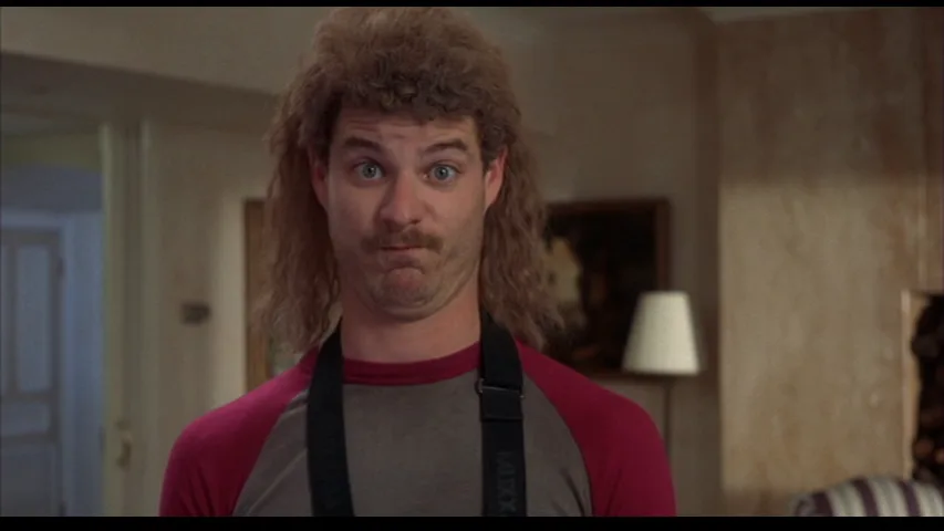 A man with a voluminous, curly mullet hairstyle and a mustache stares with wide, startled eyes. He wears a red and gray…