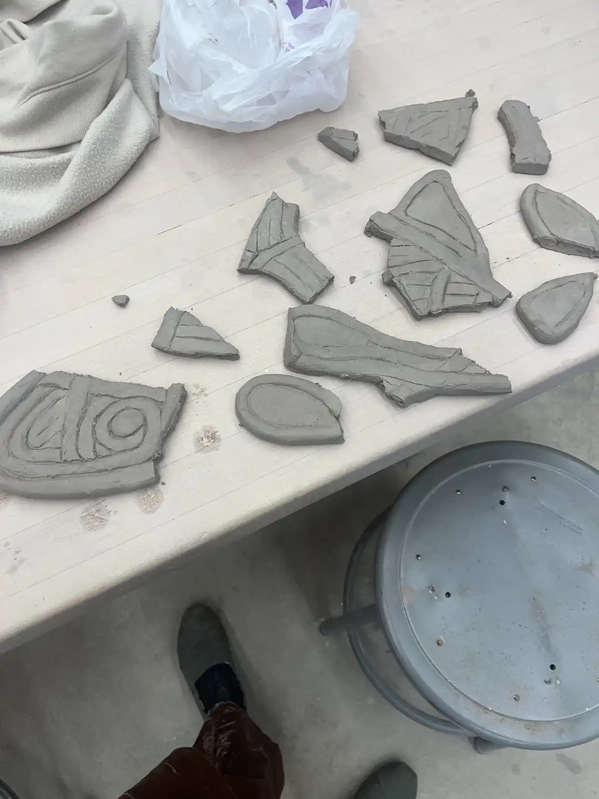 A top-down view of a cluttered pottery workbench with numerous broken pieces of grey clay scattered across its surface, some…