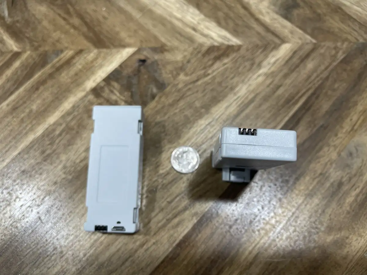 Two small, light gray electronic devices are placed on a wooden floor next to a silver coin for scale. The overlaid text…
