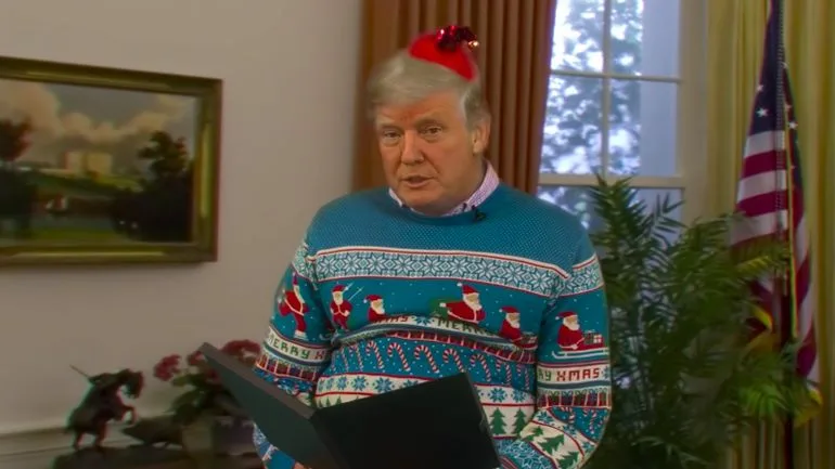 a man with gray hair wearing a blue christmas sweater with santa claus patterns and a red santa hat is holding a black…