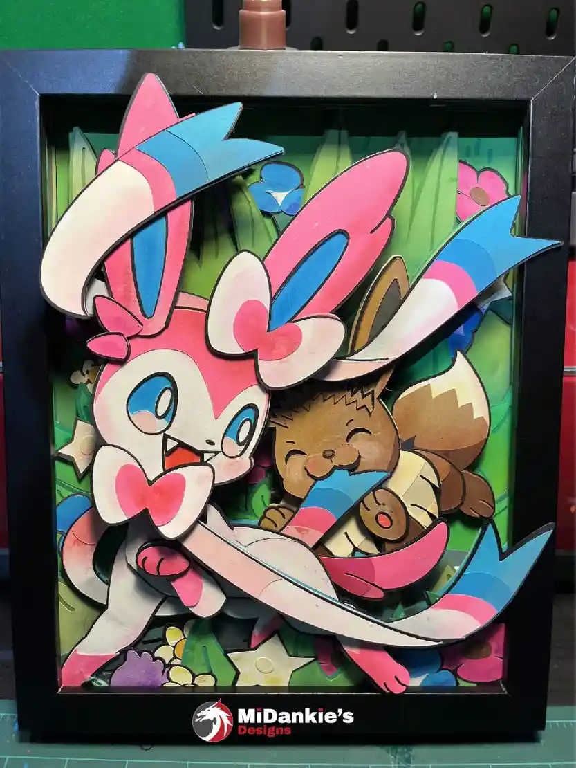 A 3D shadow box art piece featuring paper-cutout Pokémon Sylveon and Eevee in a playful pose, set against a colorful…