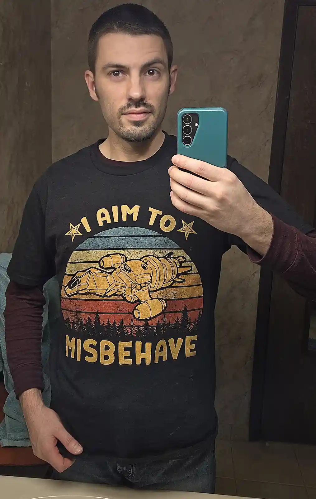 A man takes a mirror selfie holding a teal smartphone, wearing a black t-shirt with a vintage-style graphic of a yellow…