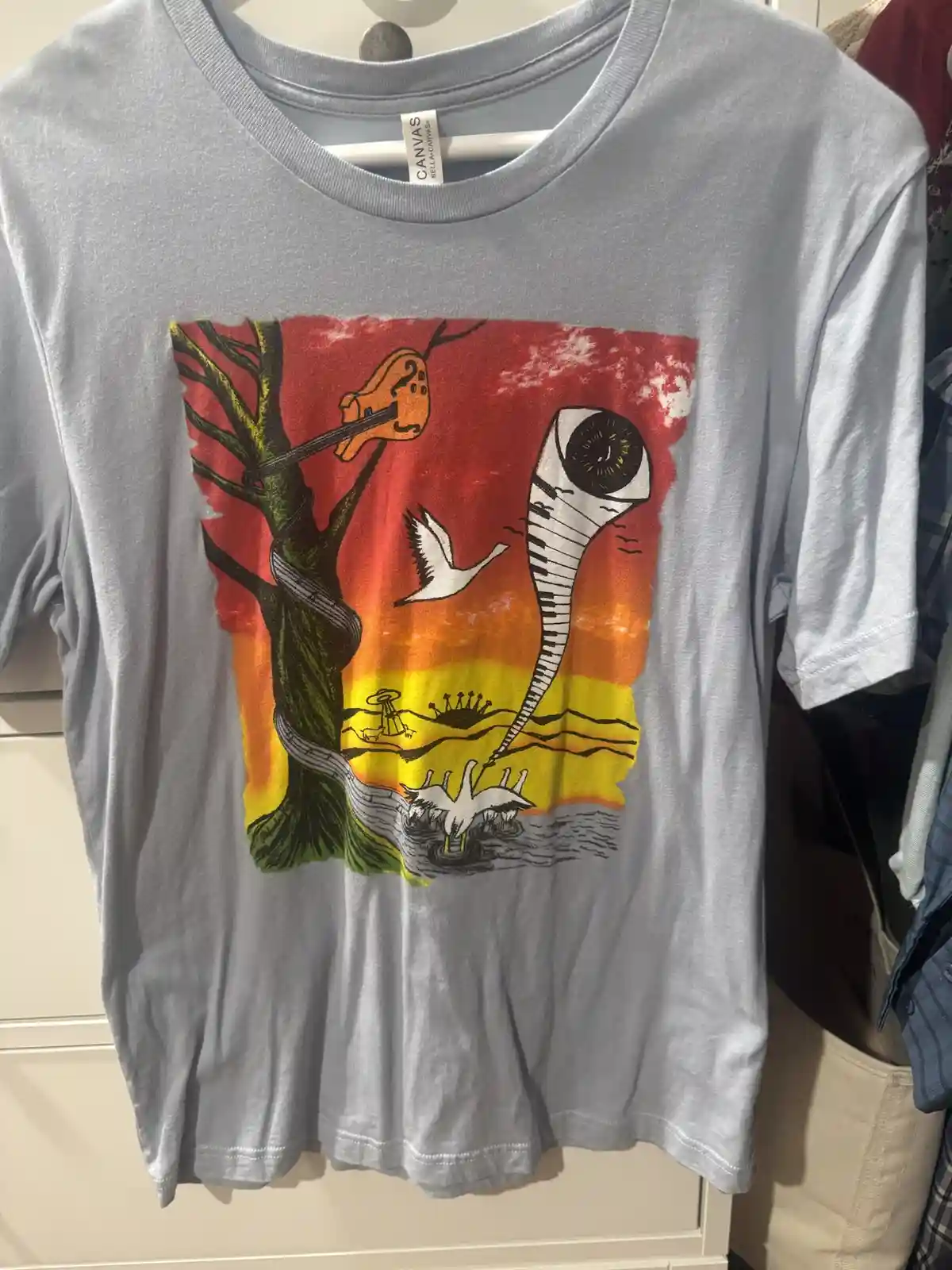 A light gray t-shirt hangs on a hanger, displaying a surreal, colorful graphic with a tree whose branches form a guitar, a…