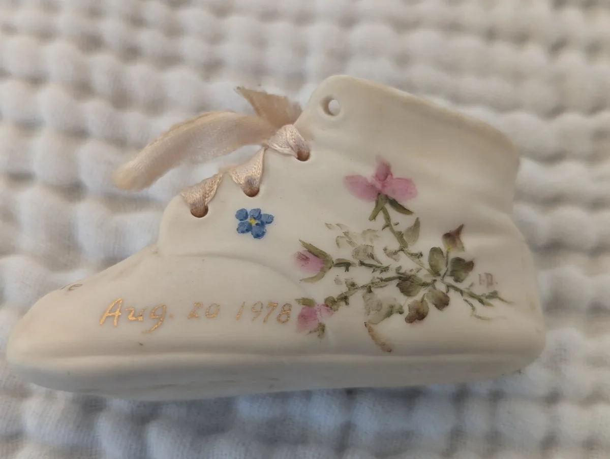 A close-up of a small, white ceramic baby shoe ornament with delicate pink and blue painted flowers and green stems. It has…