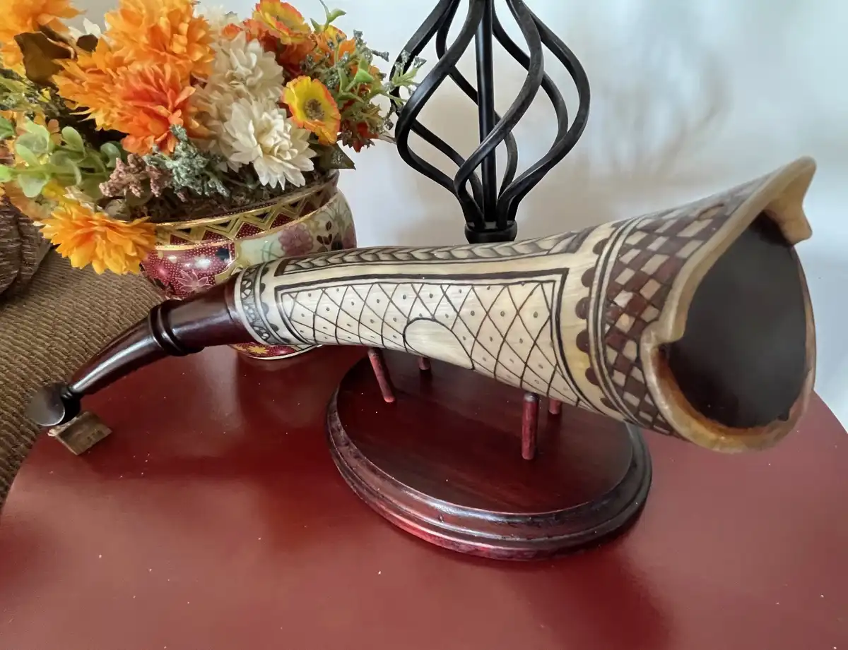 A decorative, hand-carved wooden horn with geometric patterns sits on a dark wooden stand on a red table. Behind it is a…