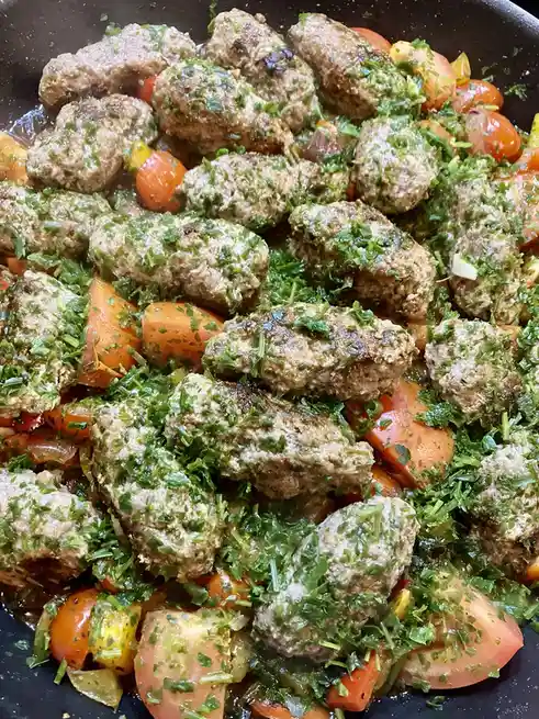 A close-up photo of a pan filled with browned meatballs, sliced tomatoes, and chopped green herbs, likely parsley, all mixed…