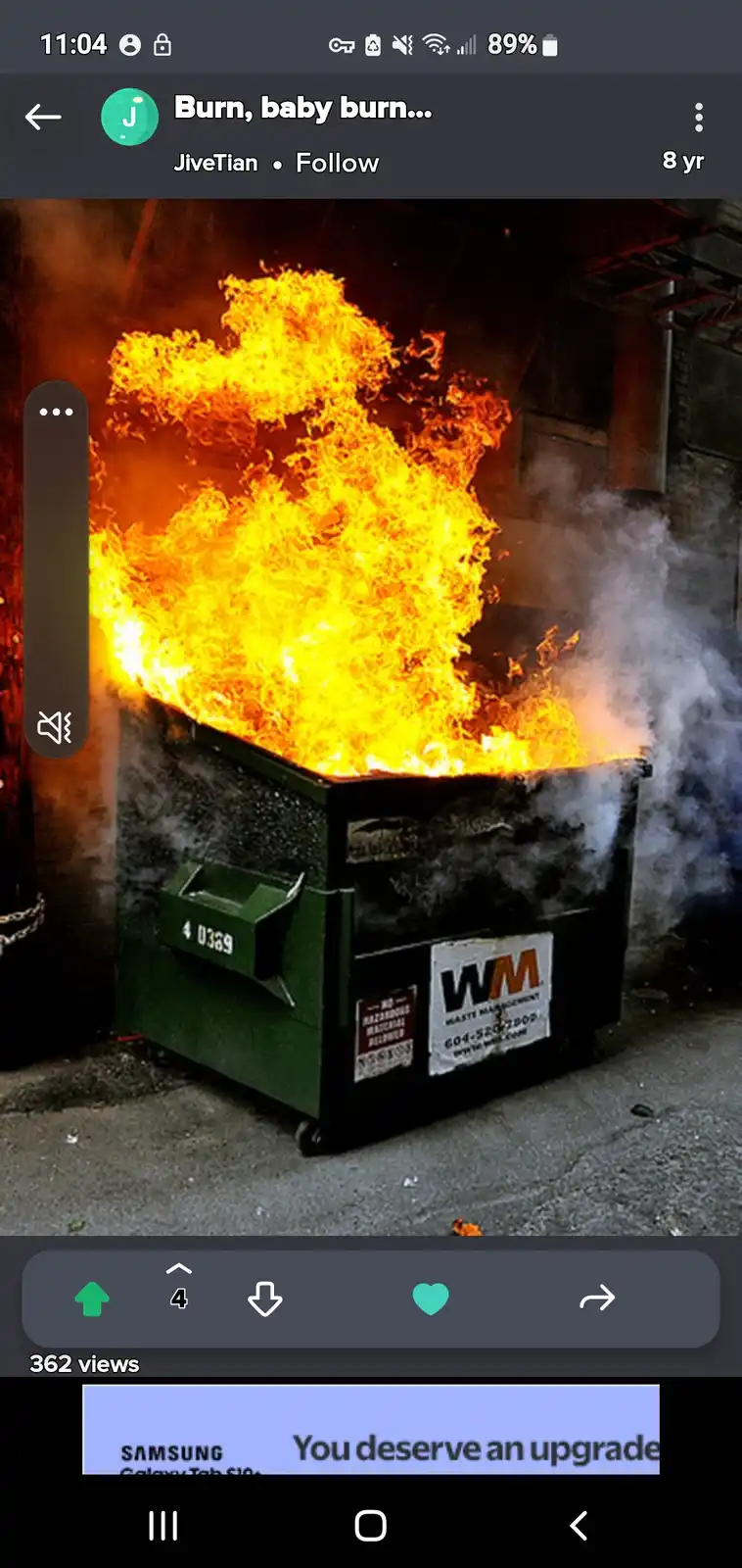 A large green dumpster with a 'WM Waste Management' sticker is engulfed in bright orange flames and billowing smoke, set…