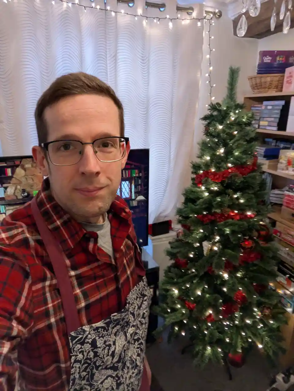 A man wearing glasses, a red plaid shirt, and a patterned apron stands in front of a decorated Christmas tree with lights…