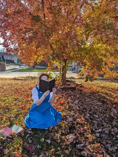 A person in a blue dress sits on grass covered in fallen autumn leaves under a tree with vibrant orange and red foliage…