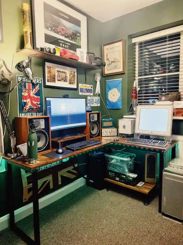 A corner desk setup features a modern flat-screen monitor next to a vintage Apple CRT monitor, both with keyboards and mice…