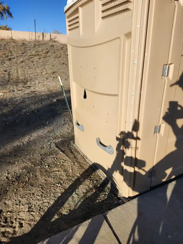 A tan portable toilet sits outdoors on gravel next to a concrete sidewalk under a clear blue sky. The side of the unit has a…