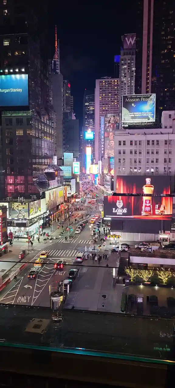 A nighttime photo of Times Square, New York, viewed from a high window with a glass of water in the foreground. The scene is…