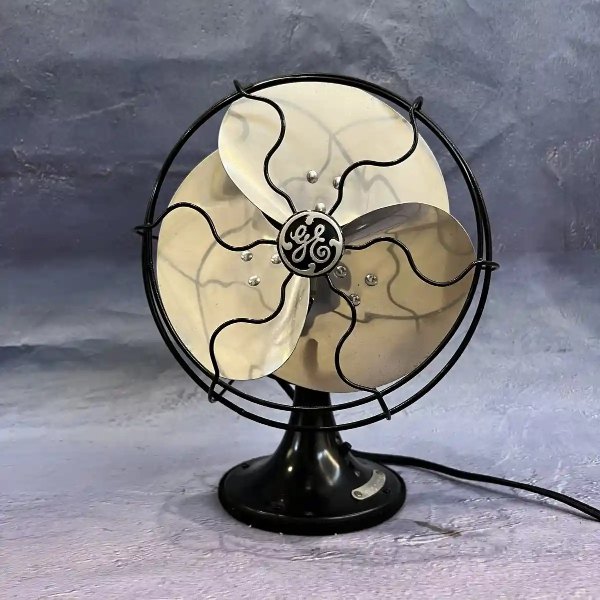 A vintage General Electric table fan with a black metal cage and brass-colored blades sits on a textured gray surface. The…