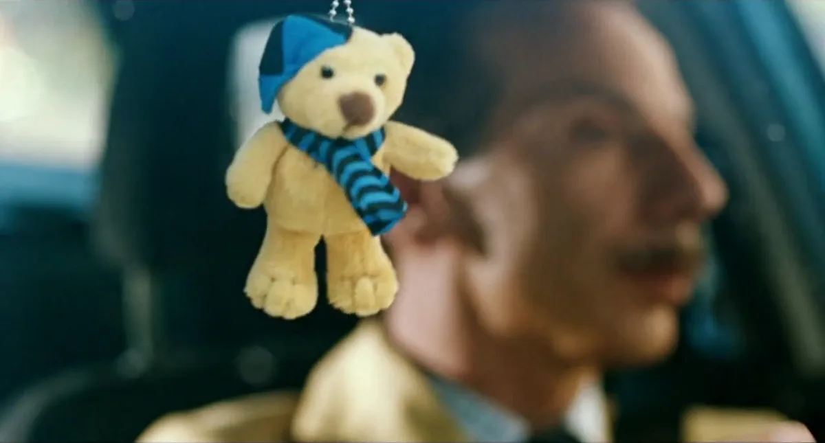 A small, light-brown plush teddy bear wearing a blue and black checkered hat and a blue and black striped scarf hangs in…