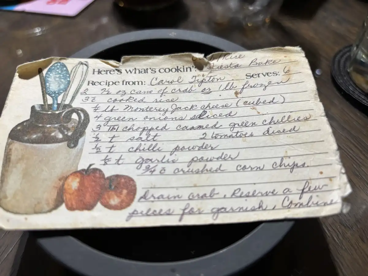 A close-up of a handwritten recipe card on aged paper, placed on a dark wooden table. The card features a printed…