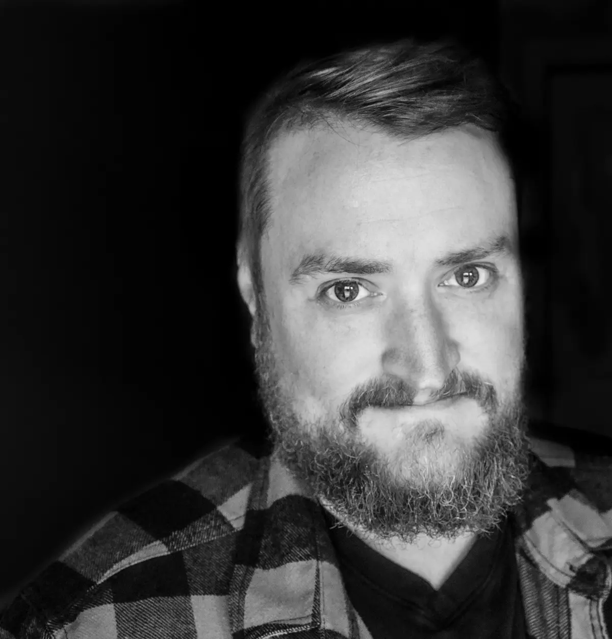 A black and white close-up photo of a man with a beard and plaid shirt looking directly at the camera with a slightly smug…