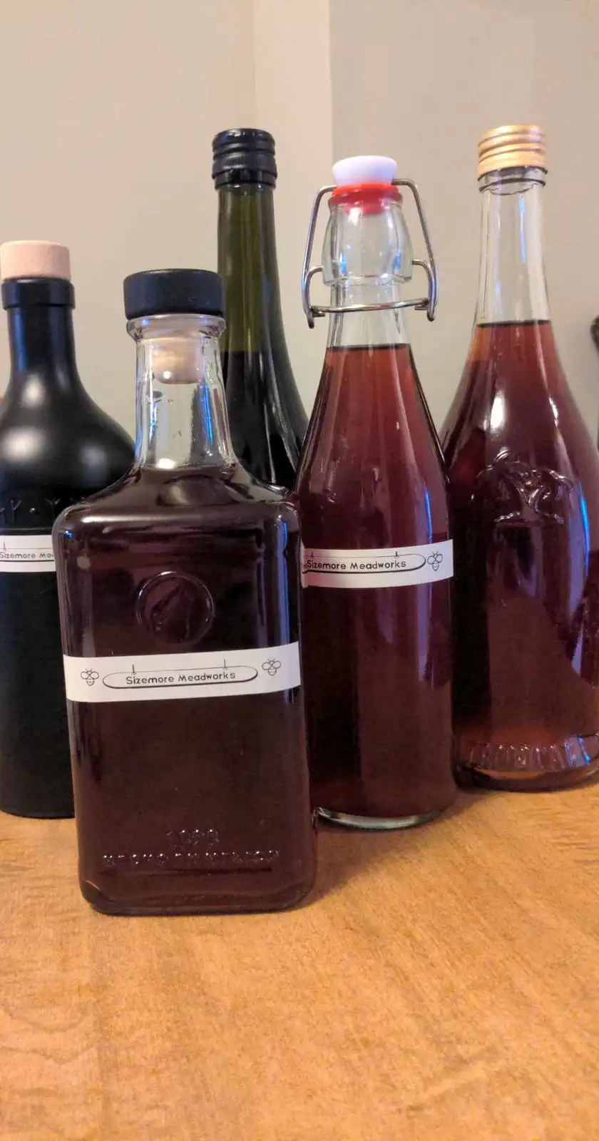 Several glass bottles filled with dark amber liquid, labeled 'Sizemore Meadworks' with bee icons, are arranged on a wooden…