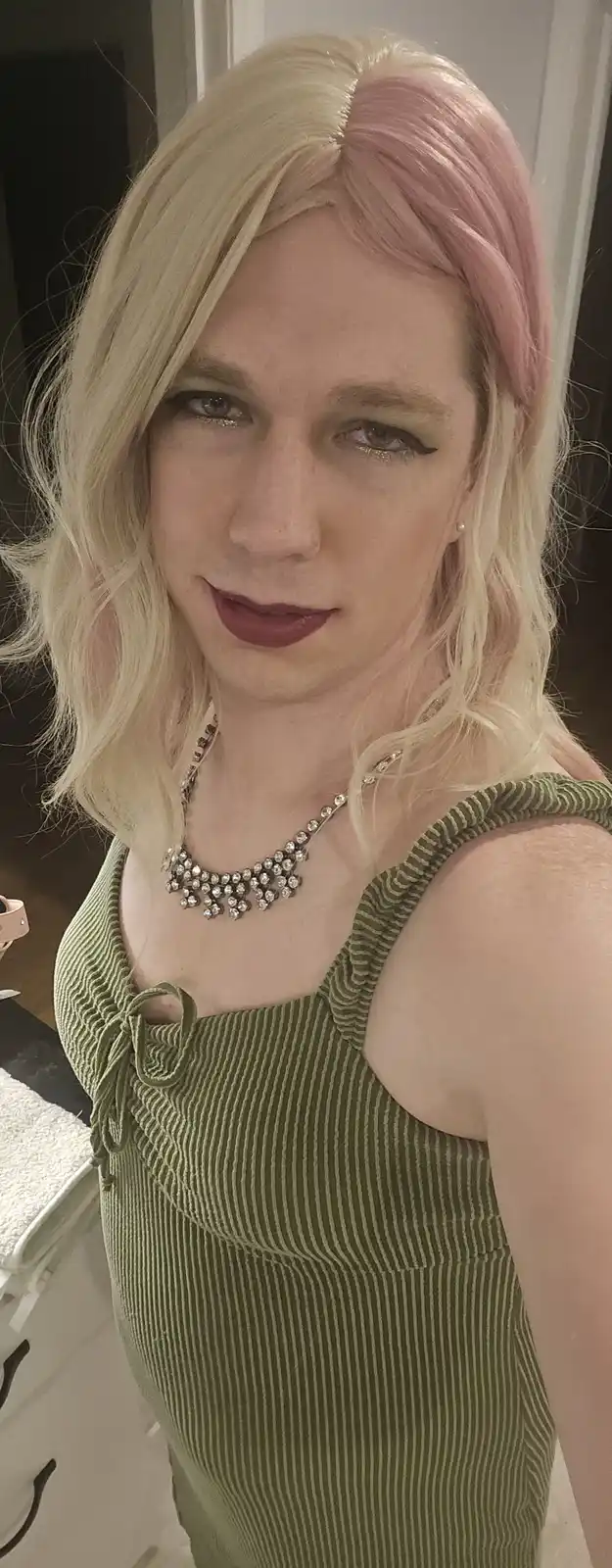 A person with blonde hair split into two colors, one side blonde and the other pink, wearing a green striped top and a…