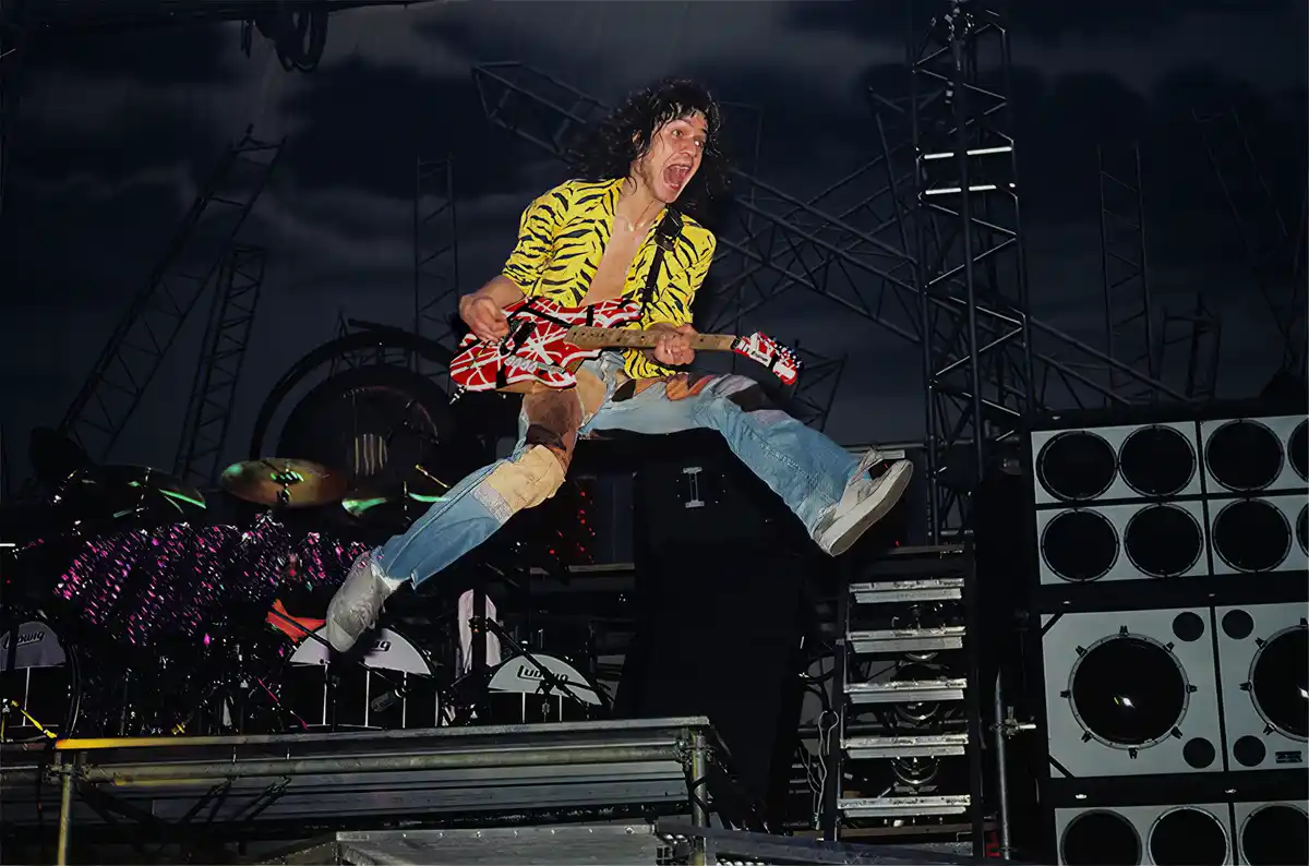 A man with long dark hair, wearing a yellow tiger-print shirt and ripped jeans, is captured mid-air while playing a red and…