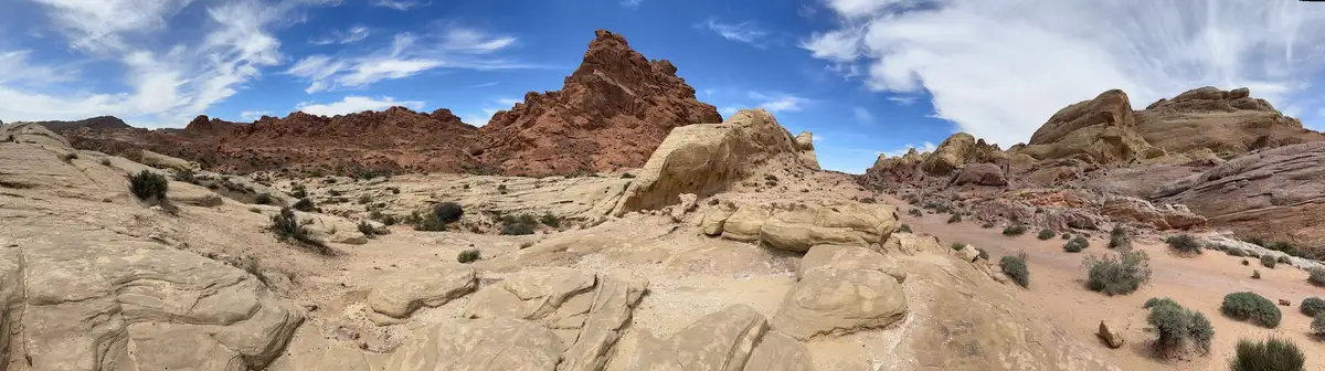 A panoramic desert landscape with red and beige rock formations under a blue sky with wispy clouds. Sparse green shrubs dot…