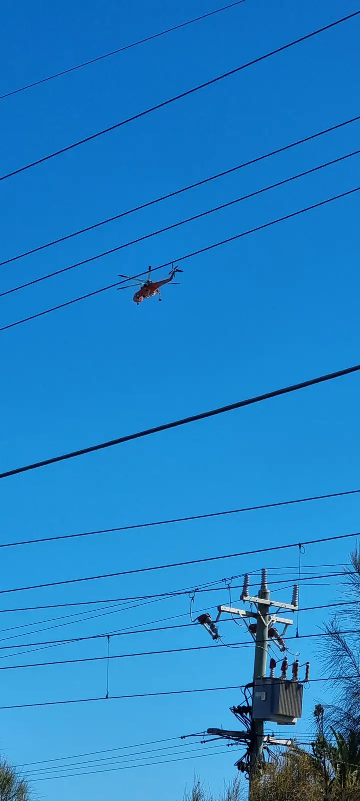 An orange helicopter is visibly hovering and seemingly stuck between multiple black power lines against a clear blue sky…