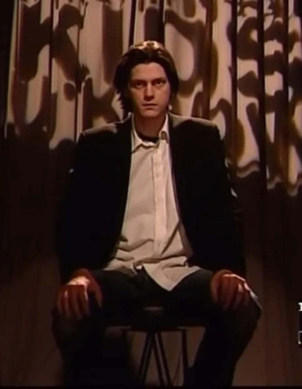 A man with dark hair, wearing a black blazer over a white button-up shirt and dark pants, sits on a stool with his hands…