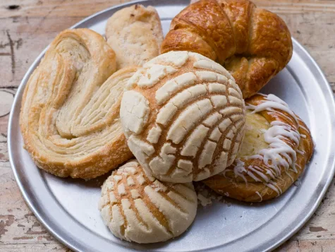 A silver plate holds assorted pastries including a croissant, palmiers, and conchas on a wooden surface. Overlaid text in…
