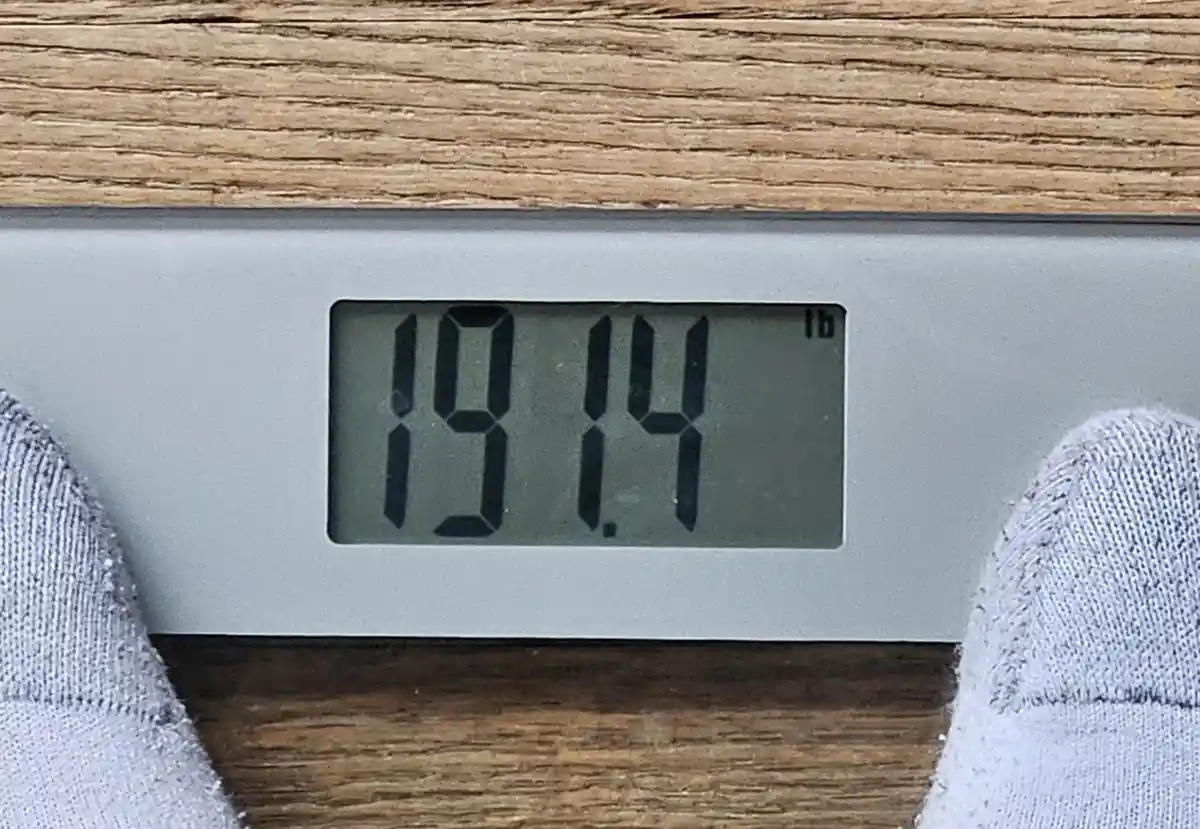 A close-up of a digital bathroom scale displaying '19.14 lb' in black digits on a gray screen, with the toes of someone…