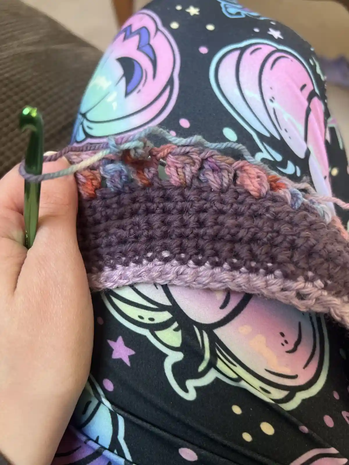A close-up photo shows a person's hand holding a green crochet hook while working on a partially completed, multicolored…